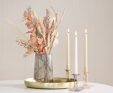 Sklenená váza s umelými kvetmi na zlatom podnose a sviečky vo farebných svietnikoch 