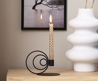 Sviečka postavená na kovovom čiernom svietniku vedľa bielej vázy