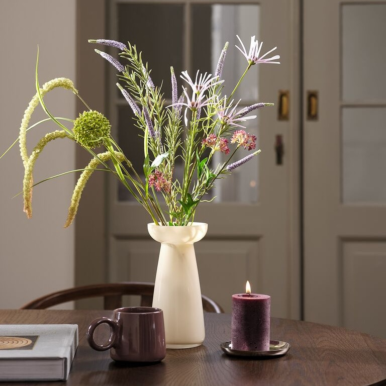 Artificial flowers White ceramic vase with artificial wildflowers in purple, pink, and green tones, on a dark wood table beside a mug and a lit candle.