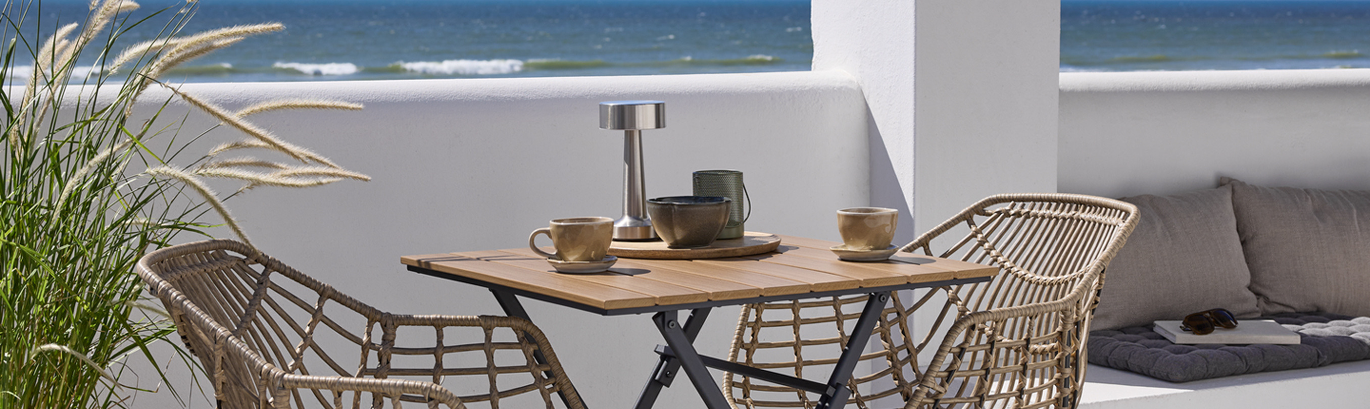 Balcony cover Balcony with a small wooden bistro table and two woven rattan chairs set with cups and a table lamp, overlooking the ocean.