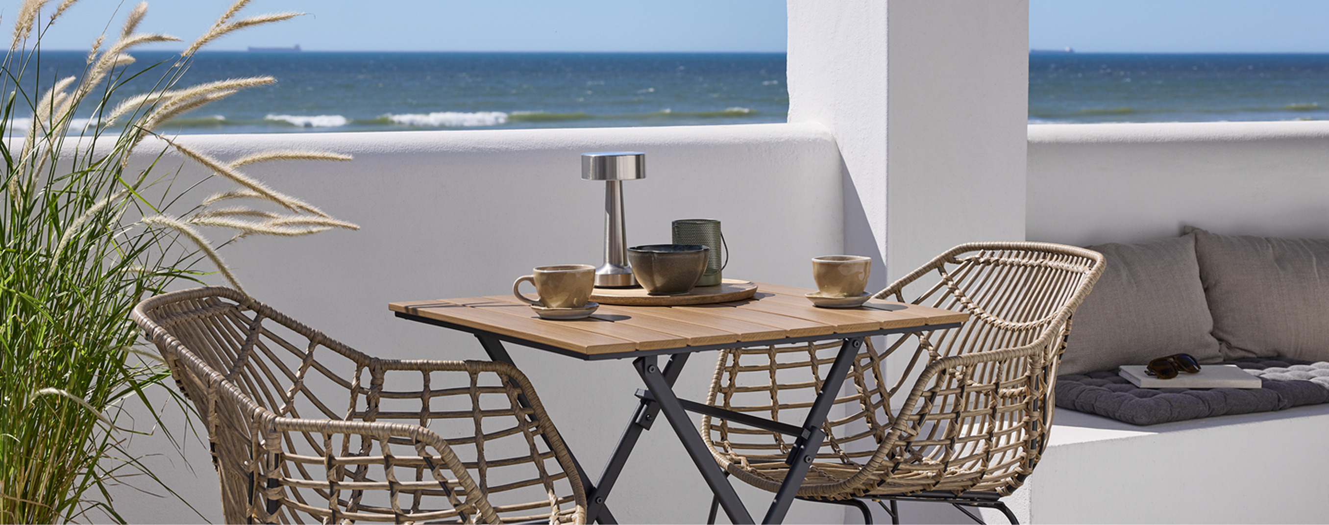 Balcony cover Balcony with a small wooden bistro table and two woven rattan chairs set with cups and a table lamp, overlooking the ocean.