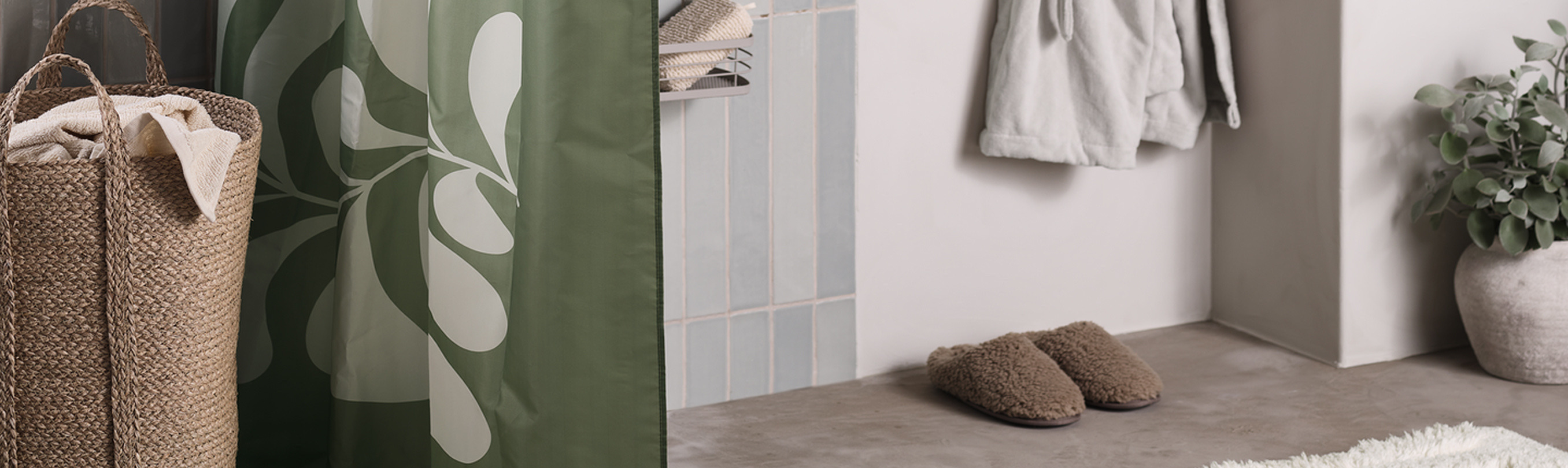 Bathroom nook with a green leaf-patterned shower curtain beside a woven jute hamper, cozy slippers, and a hanging robe.
