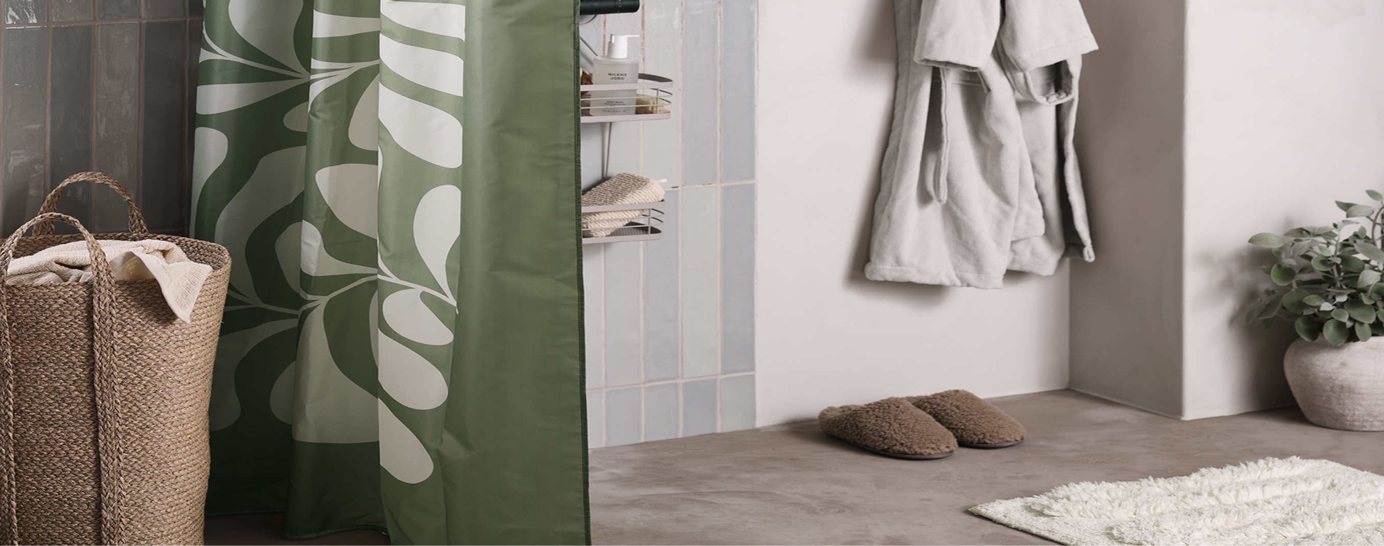 Bathroom nook with a green leaf-patterned shower curtain beside a woven jute hamper, cozy slippers, and a hanging robe.