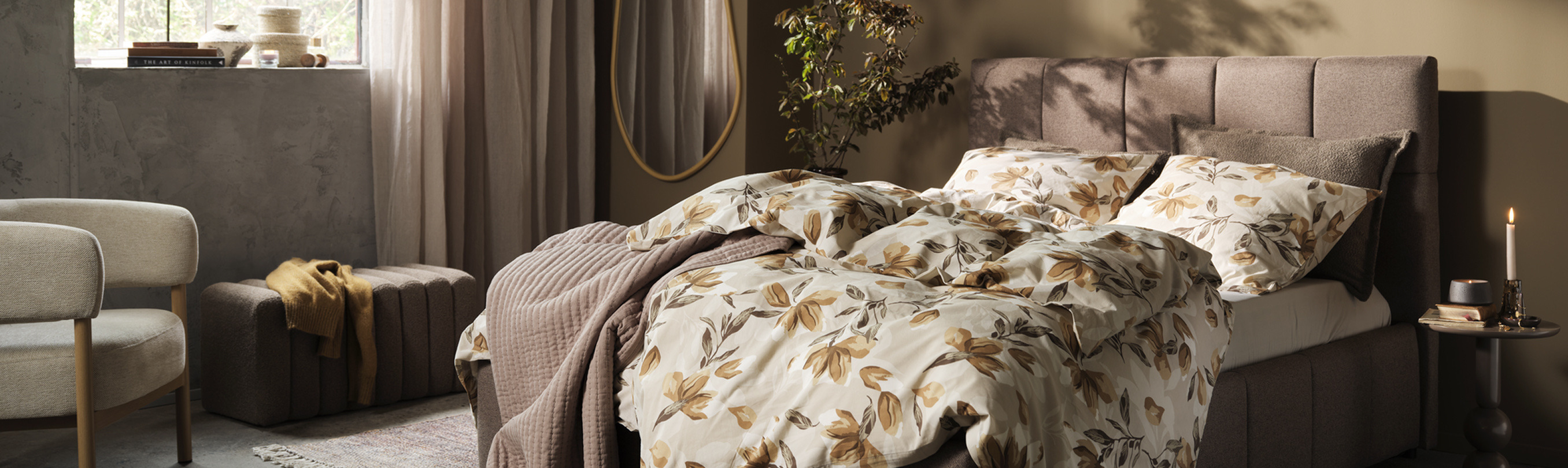 Bedroom with a taupe upholstered bed dressed in a cream and rust floral duvet, a beige armchair, a round mirror, and a potted plant, lit by warm ambient light.