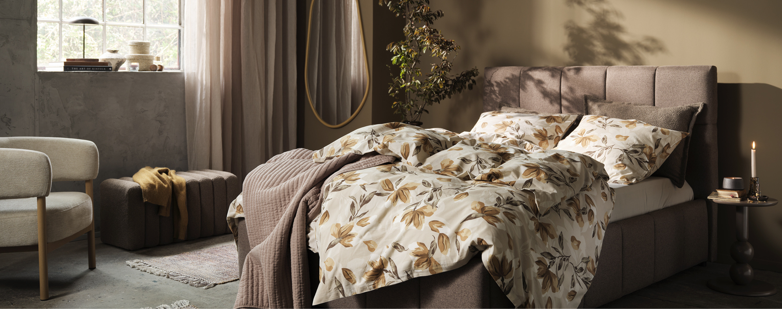 Bedroom with a taupe upholstered bed dressed in a cream and rust floral duvet, a beige armchair, a round mirror, and a potted plant, lit by warm ambient light.