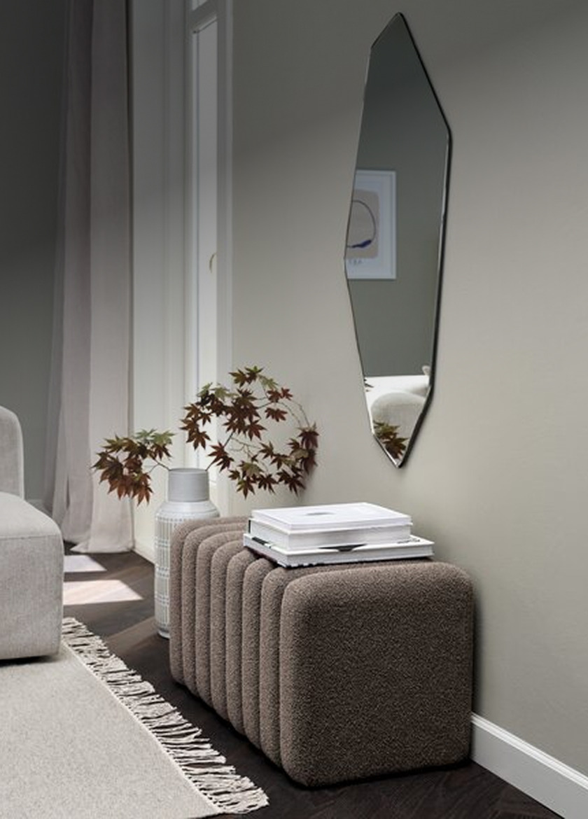 Ribbed brown bouclé bench beneath an angular wall mirror in a hallway, styled with a ceramic vase of dried leaves and stacked books.
