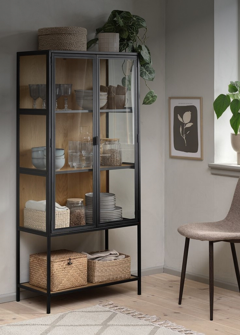 Black-framed glass display cabinet with wood-backed shelves holding glassware, bowls, and jars, beside a potted plant and framed art.