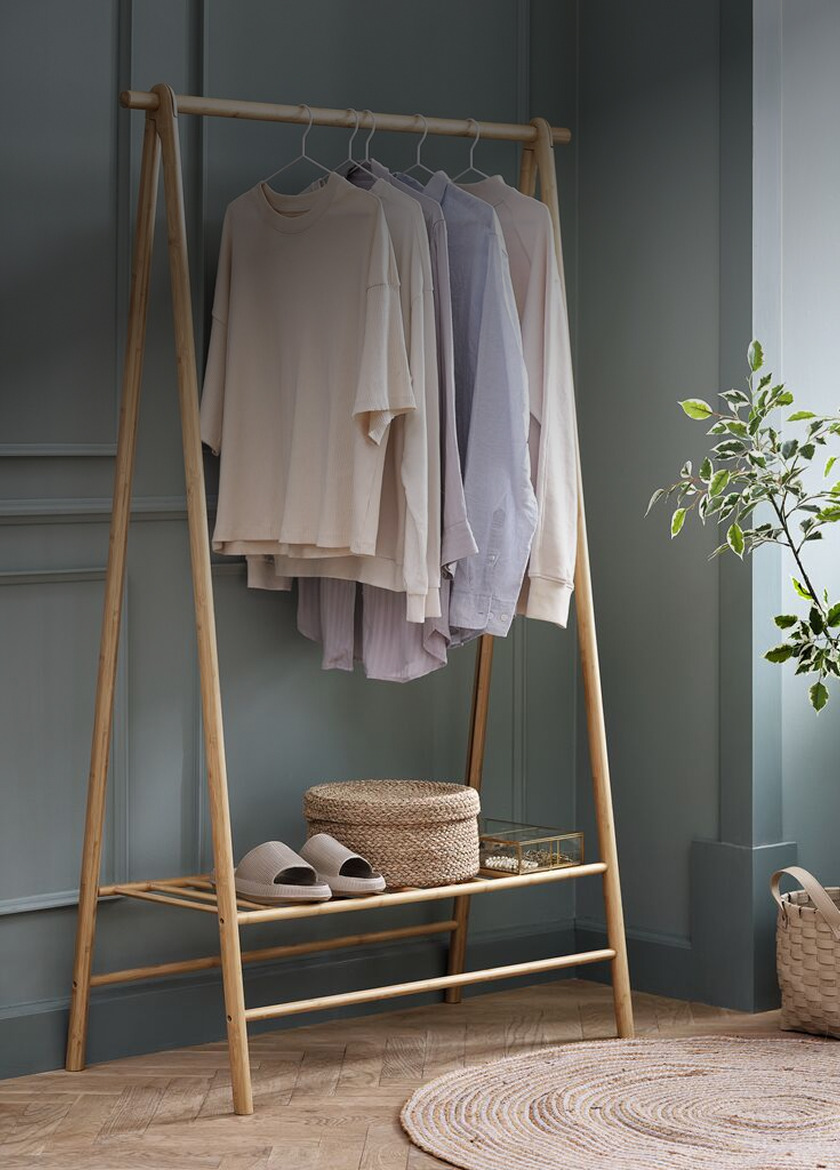 Freestanding wooden A-frame clothes rail with folded sweaters and shirts on hangers, slippers and a woven basket on the lower shelf, beside a potted plant.