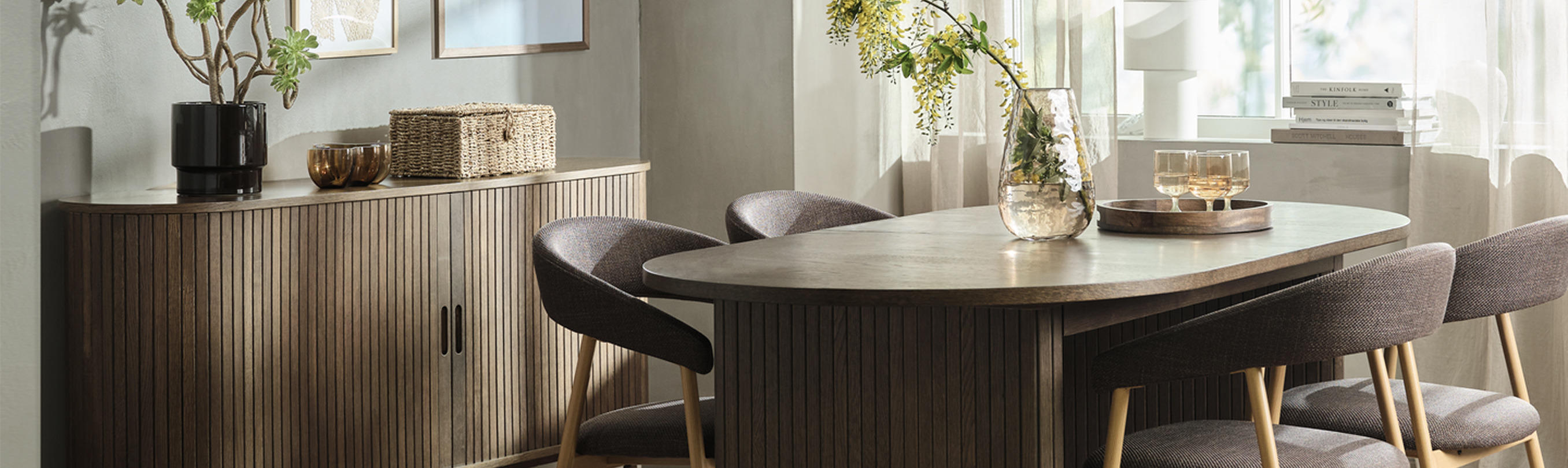Dining room with a ribbed wood sideboard, framed art, and an oval wood table surrounded by upholstered chairs, with a glass vase of flowering branches.