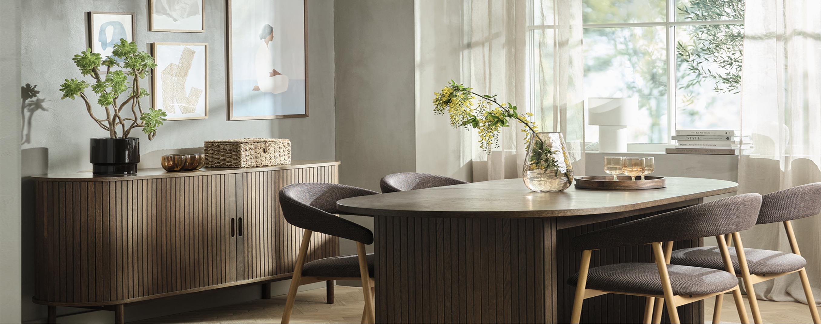 Dining room with a ribbed wood sideboard, framed art, and an oval wood table surrounded by upholstered chairs, with a glass vase of flowering branches.