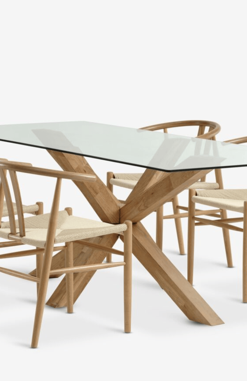 Close-up of a glass-top dining table with a crossed oak leg base surrounded by wishbone-style dining chairs with woven seats.