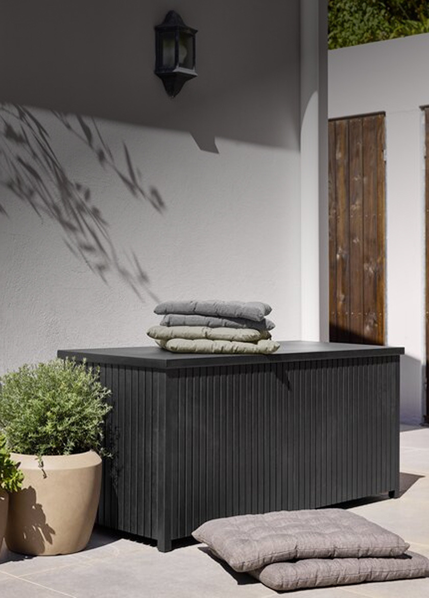 Durable storage Black ridged outdoor storage box topped with folded grey and green cushions, beside a potted plant and additional cushions on the ground.