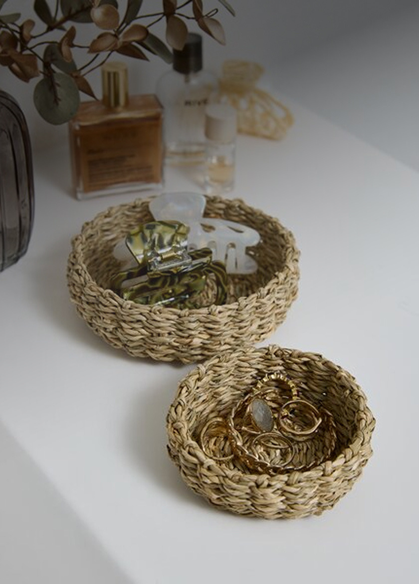 Two woven seagrass trinket bowls holding hair clips and gold jewelry, beside perfume bottles on a white shelf.