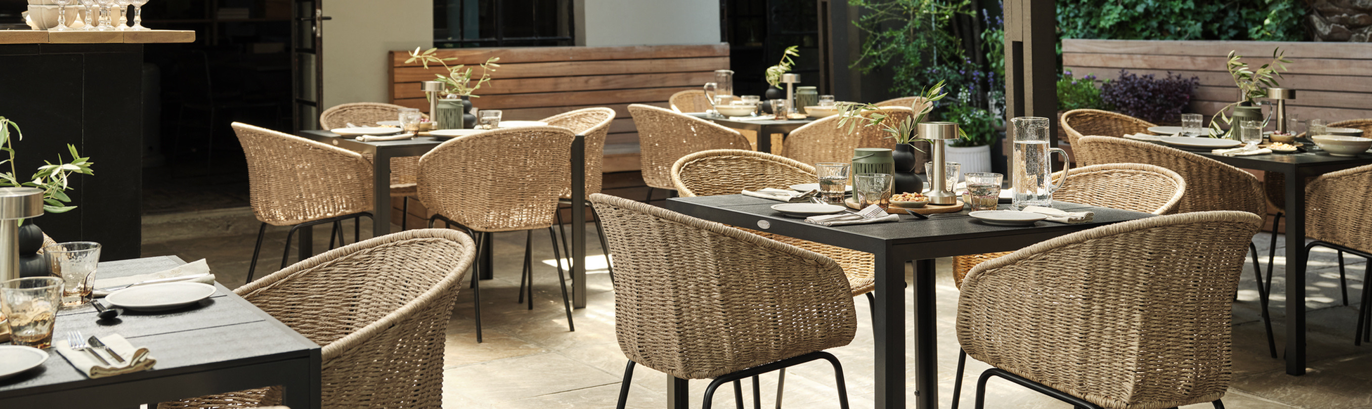 Garden cover Outdoor garden dining terrace with wicker chairs around dark tables set for a meal, surrounded by greenery and warm lighting.