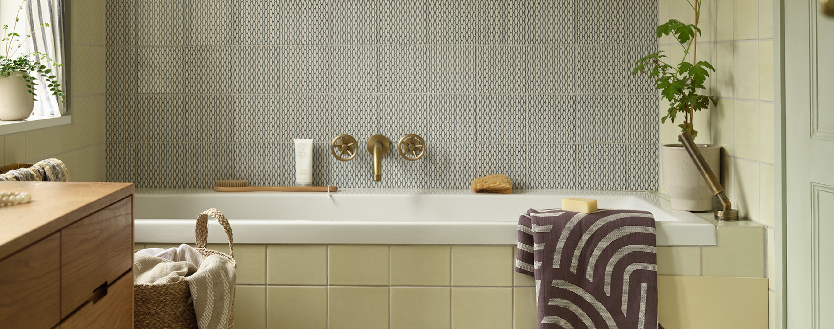 Laundry room with a patterned tile accent wall, brass fixtures, a bathtub, a woven basket, and a striped towel.