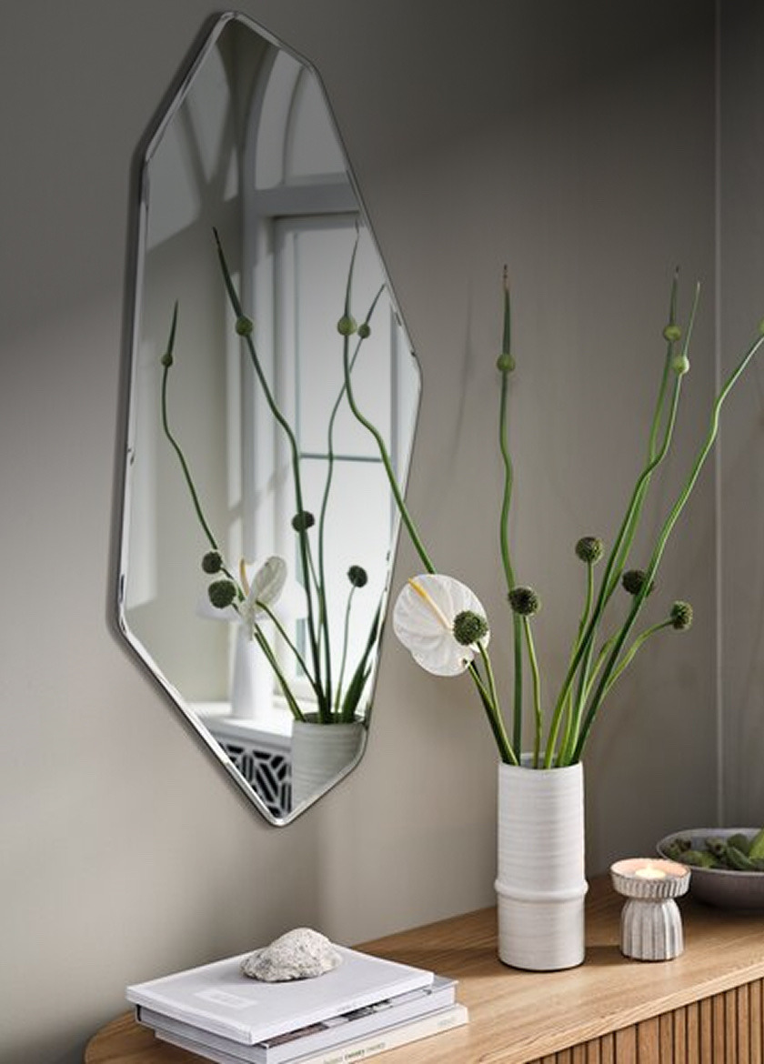 Angular diamond-shaped wall mirror above a wood shelf styled with a vase of greenery, a candle, and books.