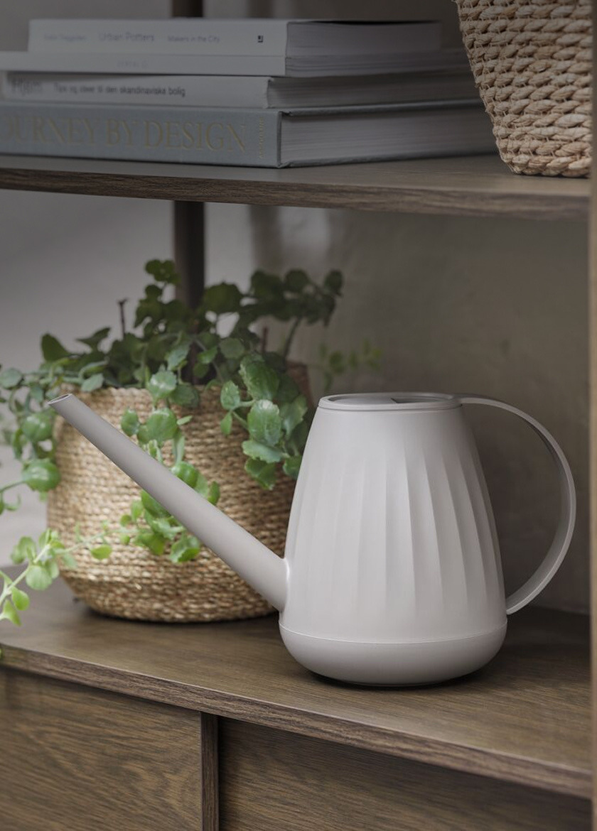 Grey watering can with a long spout, beside a woven basket planter and a stack of books on a wood shelf.