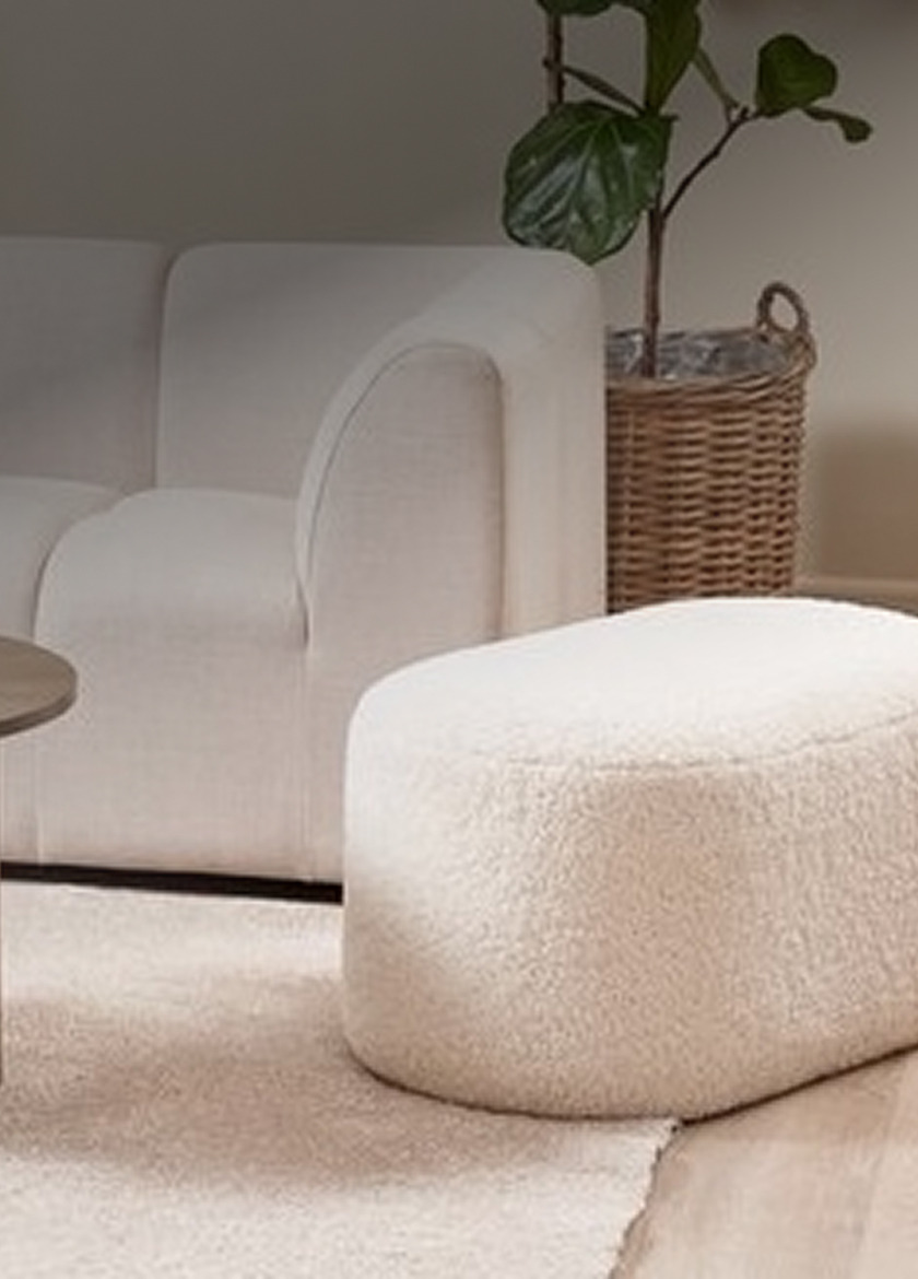 Cream bouclé oval pouffe beside a woven rattan planter and a white sofa in a living room.
