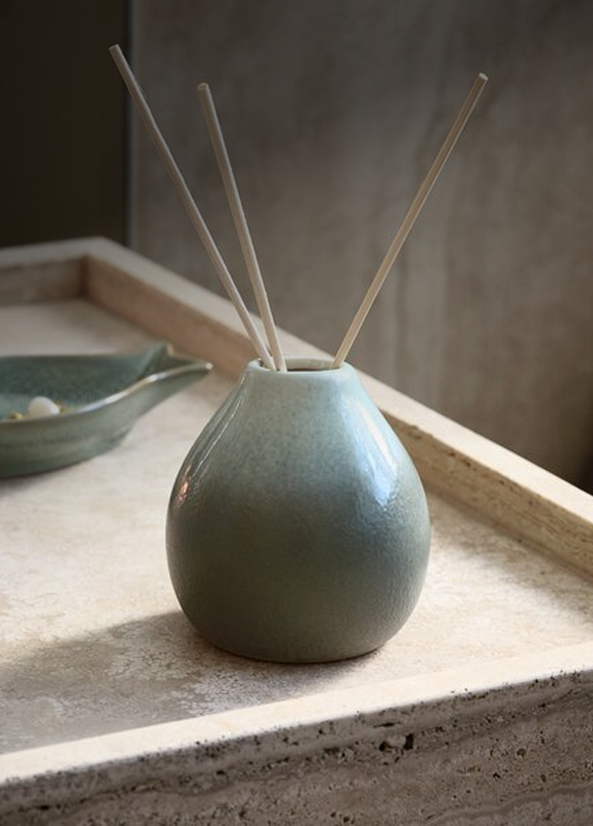 Teal ceramic reed diffuser with wooden reed sticks, on a stone tray beside a small dish, in soft natural light.