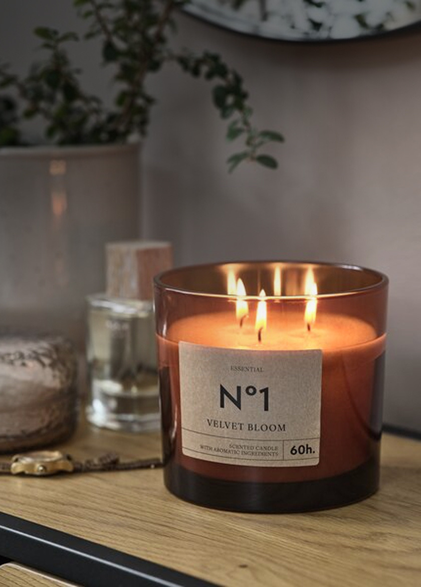 Amber glass jar candle labeled 'No1 Velvet Bloom' with three lit wicks, on a wood shelf beside a diffuser bottle and a potted plant.