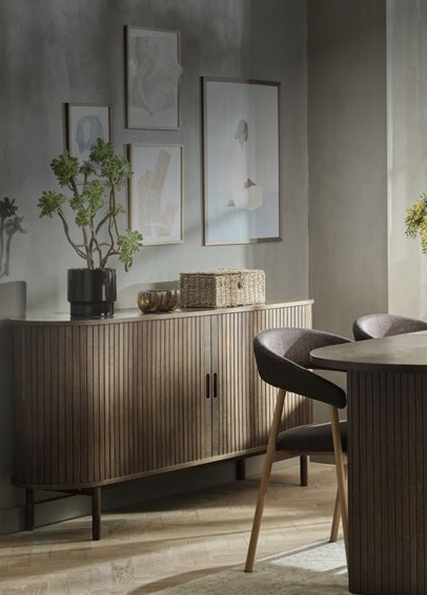 Dark wood ribbed sideboard styled with a potted plant and a woven basket, beneath framed art in a dining area.
