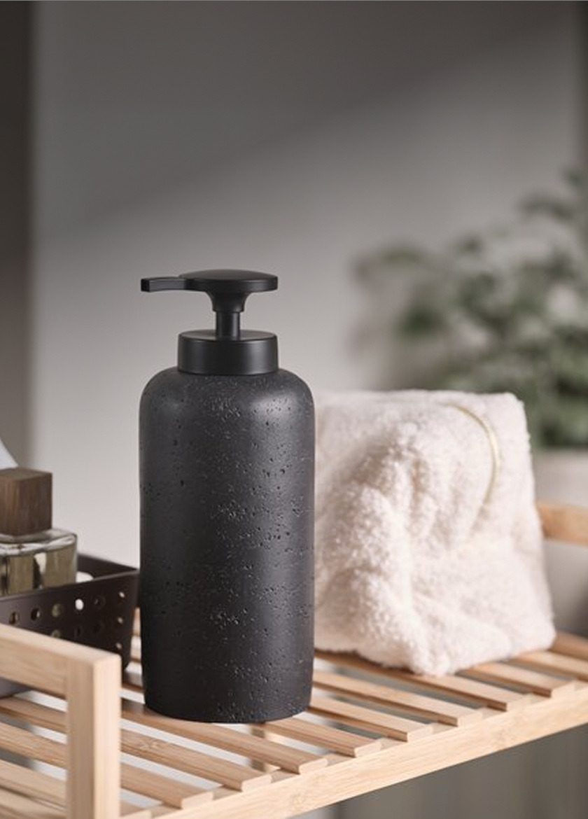 Dark textured stone soap dispenser bottle beside a folded white towel and a potted plant on a wood shelf.