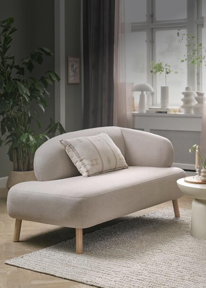 Beige upholstered two-seater sofa with a curved backrest and a striped accent pillow, in a living room with plants and a round side table.