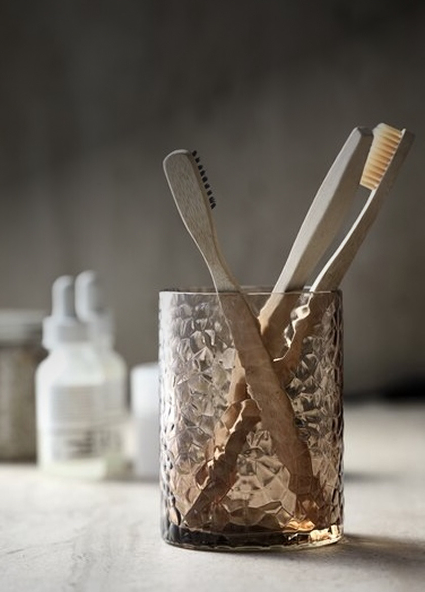 Textured glass tumbler holding two wooden toothbrushes, with toiletry bottles blurred in the background.