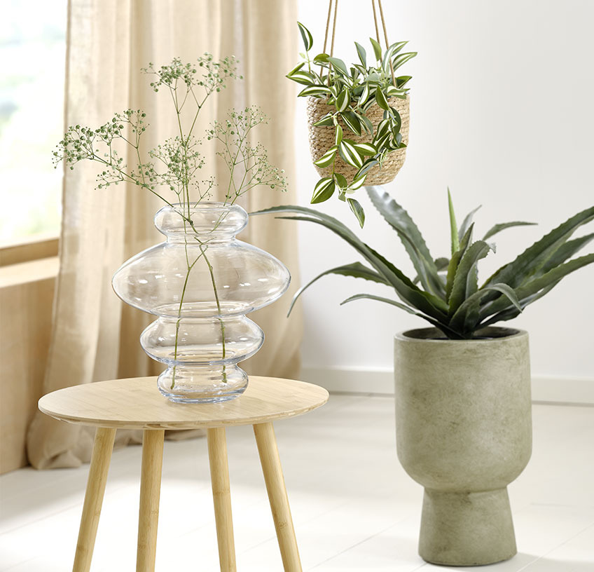 Sklenená váza na odkladacom stolíku, závesný kvetináč a zelený kvetináč s umelými rastlinami