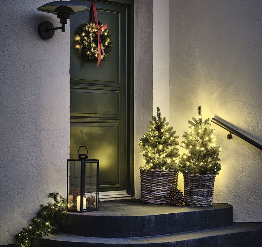 Dva svietiace stromčeky pred dverami a vianočný veniec s červenou mašľou