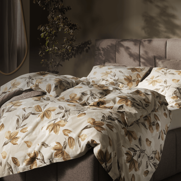 Accommodations guides Bedroom with a taupe upholstered bed dressed in a cream and rust floral duvet, a beige armchair, a round mirror, and a potted plant, lit by warm ambient light.