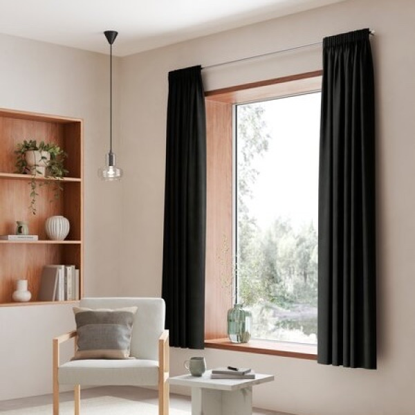 Black curtains framing a window with a view of greenery, in a room with a wood shelving unit, books, and a white armchair with a grey cushion.
