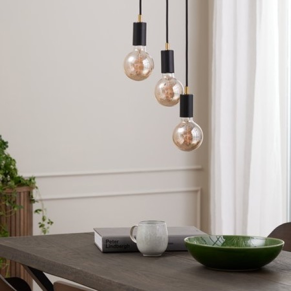 Three black pendant lights with exposed round filament bulbs hanging above a dark wood dining table set with a book, a mug, and a green ceramic bowl.