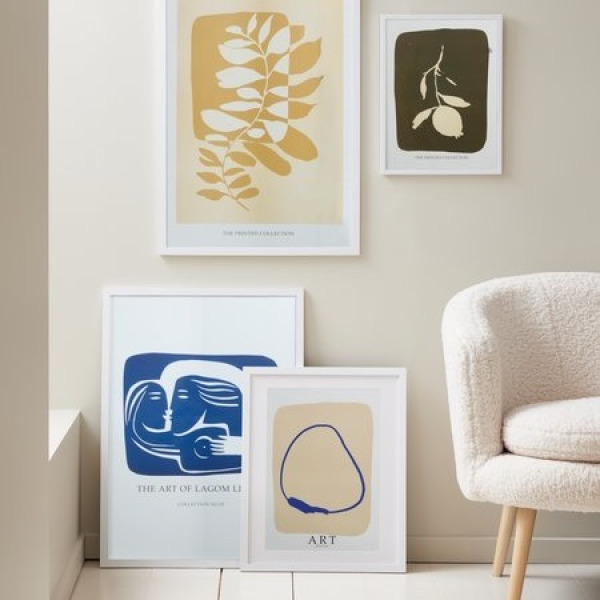 White picture frames leaning against a wall and hanging above, holding botanical and abstract art prints in yellow, blue, and olive tones, beside a cream bouclé armchair.