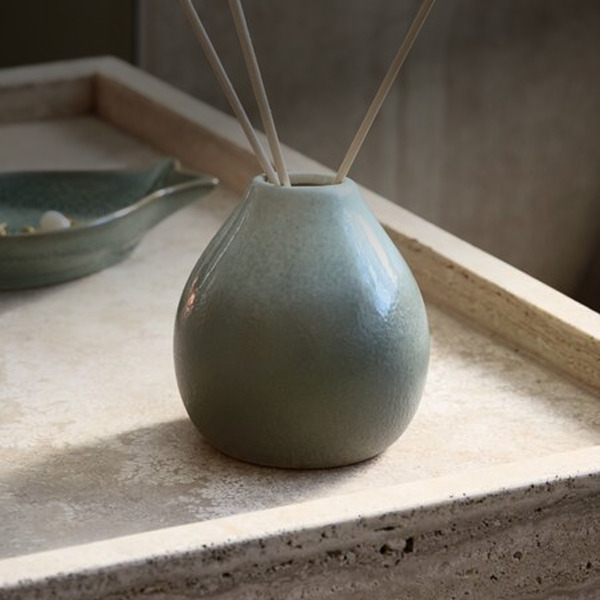 Teal ceramic reed diffuser with wooden reed sticks, on a stone tray beside a small dish.