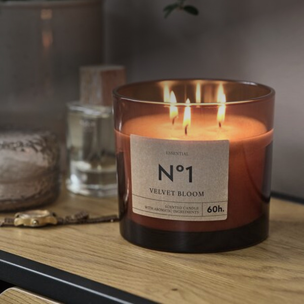 Amber glass jar candle labeled 'No1 Velvet Bloom' with three lit wicks, on a wood table beside a potted plant.