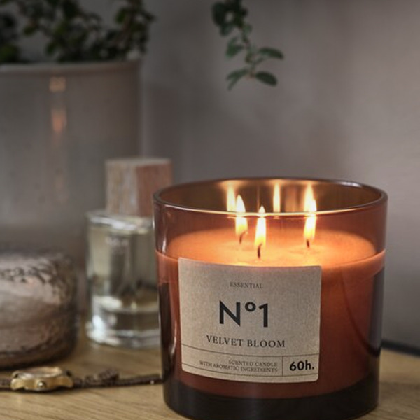 Scented candles Amber glass jar candle labeled 'No1 Velvet Bloom' with three lit wicks, on a wood shelf beside a diffuser bottle and a potted plant.