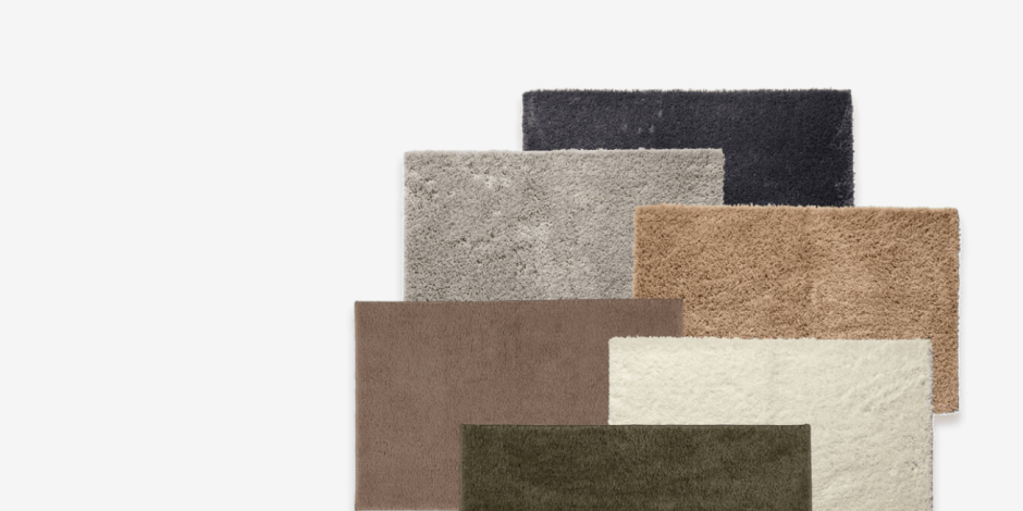 Stack of layered bath mats in black, grey, taupe, beige, cream, and olive green, shown against a plain light background.