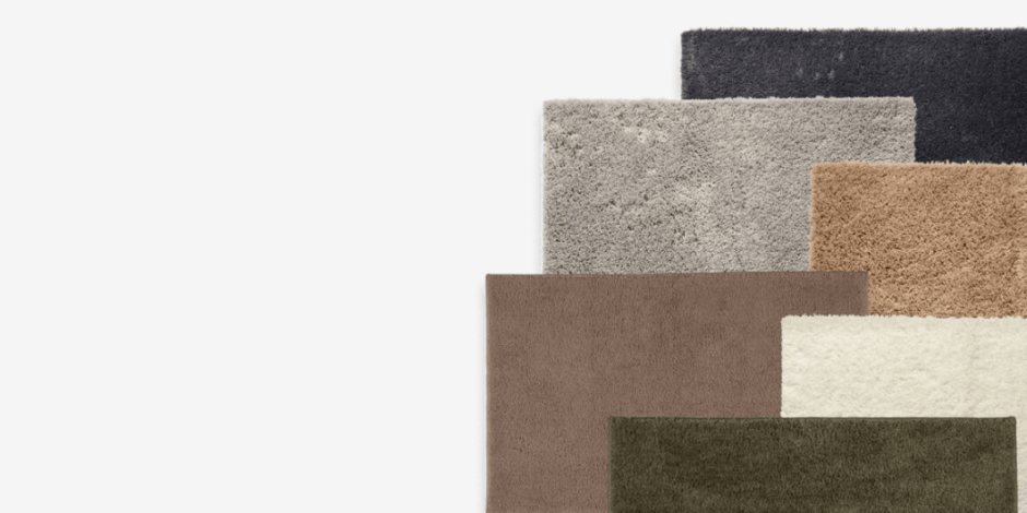 Stack of layered bath mats in black, grey, taupe, beige, cream, and olive green, shown against a plain light background.