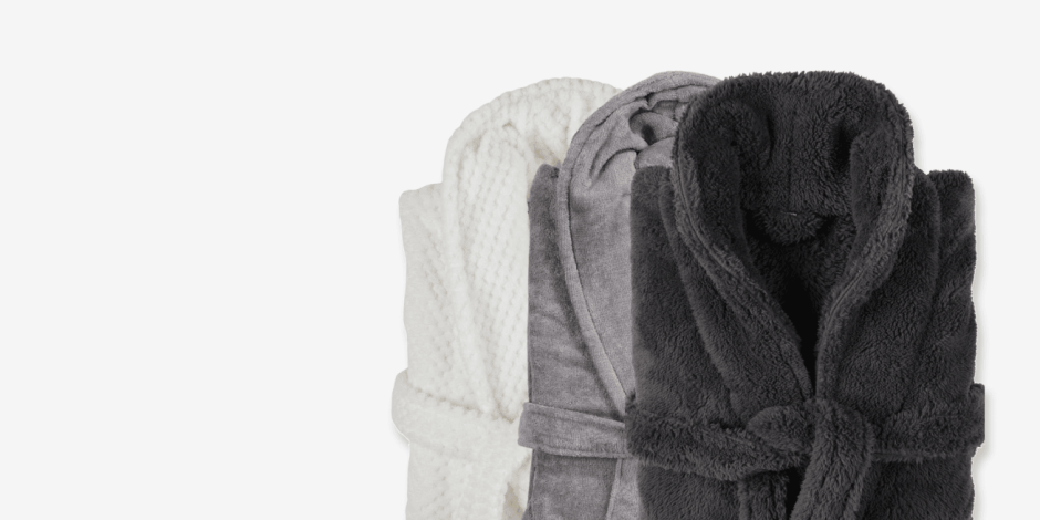 Three rolled bathrobes in white, grey, and charcoal plush fabric, shown against a plain light background.
