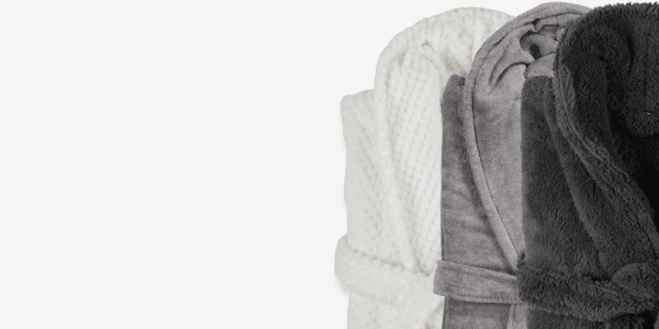 Three rolled bathrobes in white, grey, and charcoal plush fabric, shown against a plain light background.
