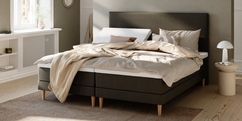Dark grey upholstered bed with beige linens, a folded knit throw, striped accent pillows, and a round bedside table with a mushroom lamp in a sunlit bedroom