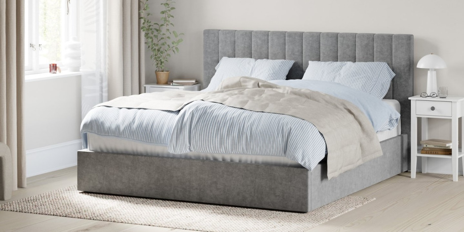 Light grey channel-tufted upholstered bed with blue and white striped bedding, a beige throw, and a white nightstand with a mushroom lamp and books.